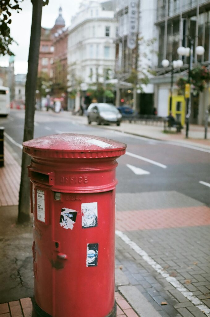 A close up of a red mail box. In the background, a blurred view of a city street with buildings and cars.