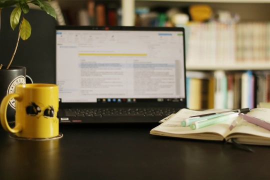 A laptoop on a desk close to a yellow mug, a little plant and a notebook with pens.